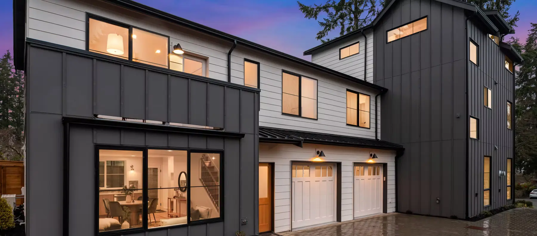 Completed dark-clad home at dusk with warm interior lighting, two carriage-style garage doors and paver drive